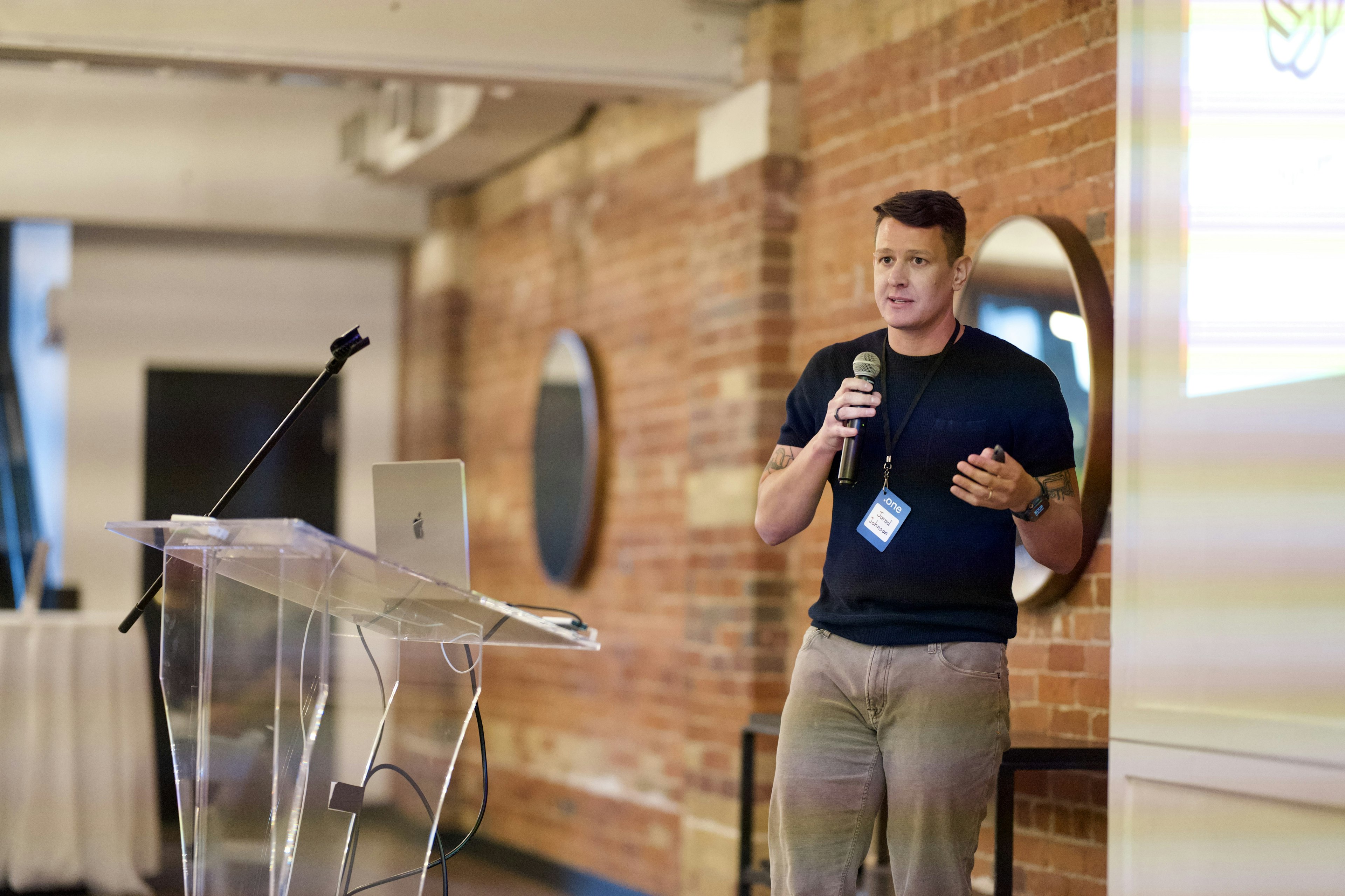 A speaker presenting at the Craft CMS Dot One conference, holding a microphone in front of projected slides.