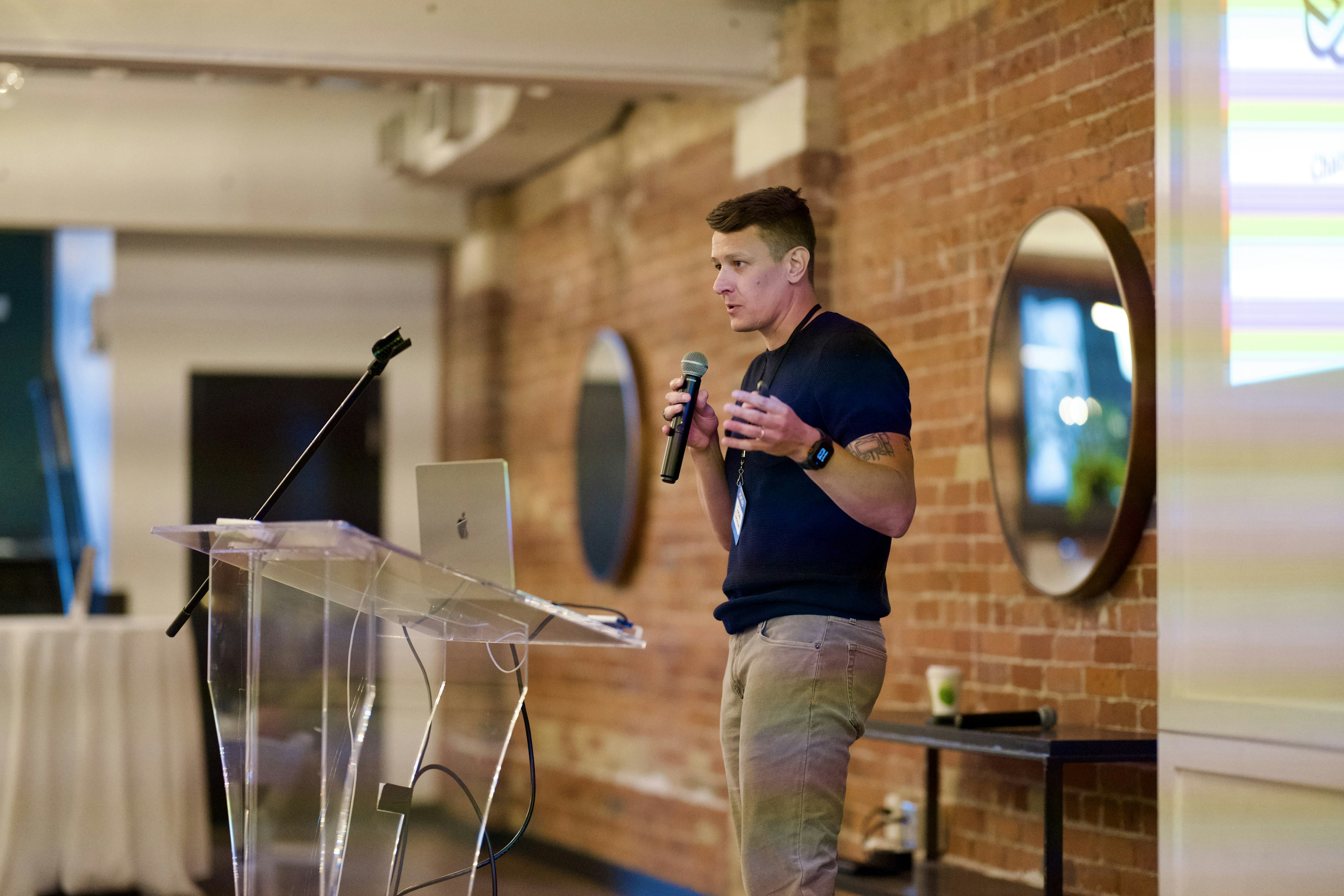 A side view of a speaker presenting at the Craft CMS Dot One conference, standing at a clear podium with a laptop.