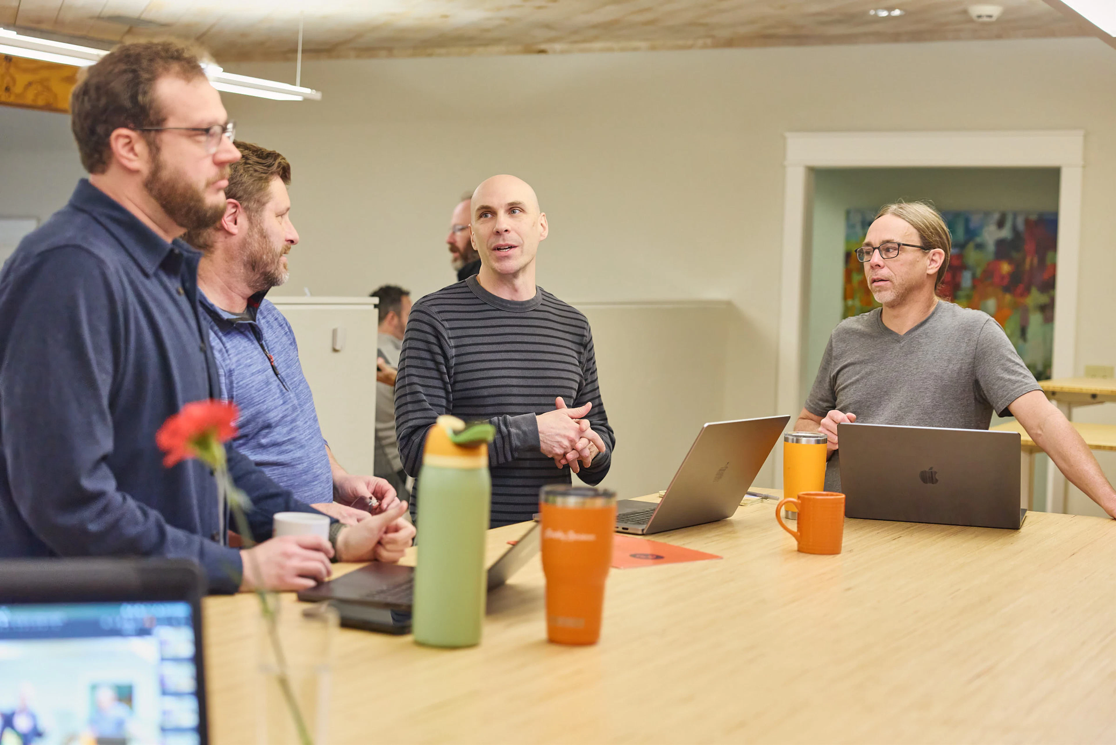 Team members standing around a table with laptops during a conversation in a bright office.
