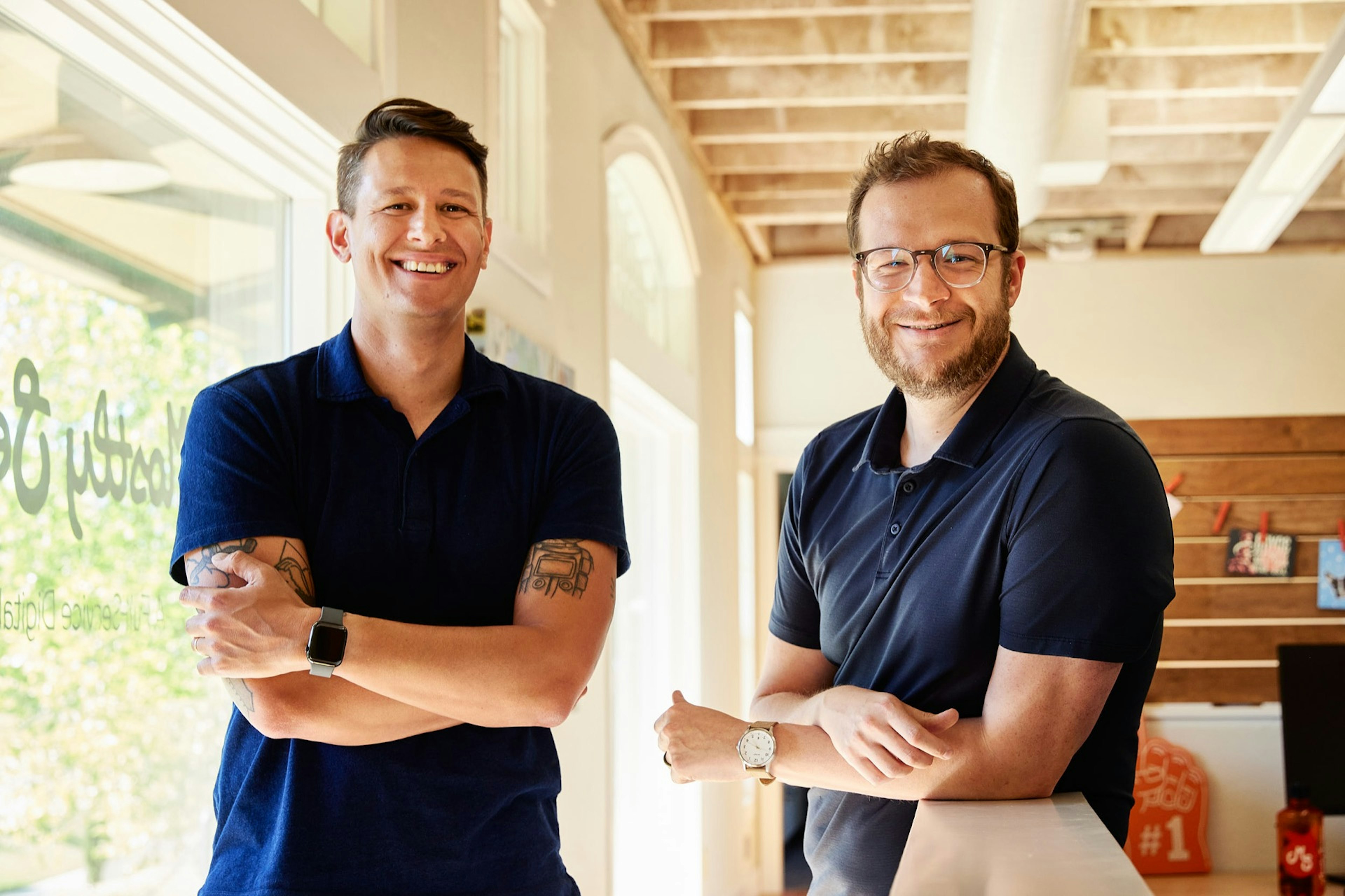 Two people standing in a bright, modern office space, smiling with arms crossed near a window.