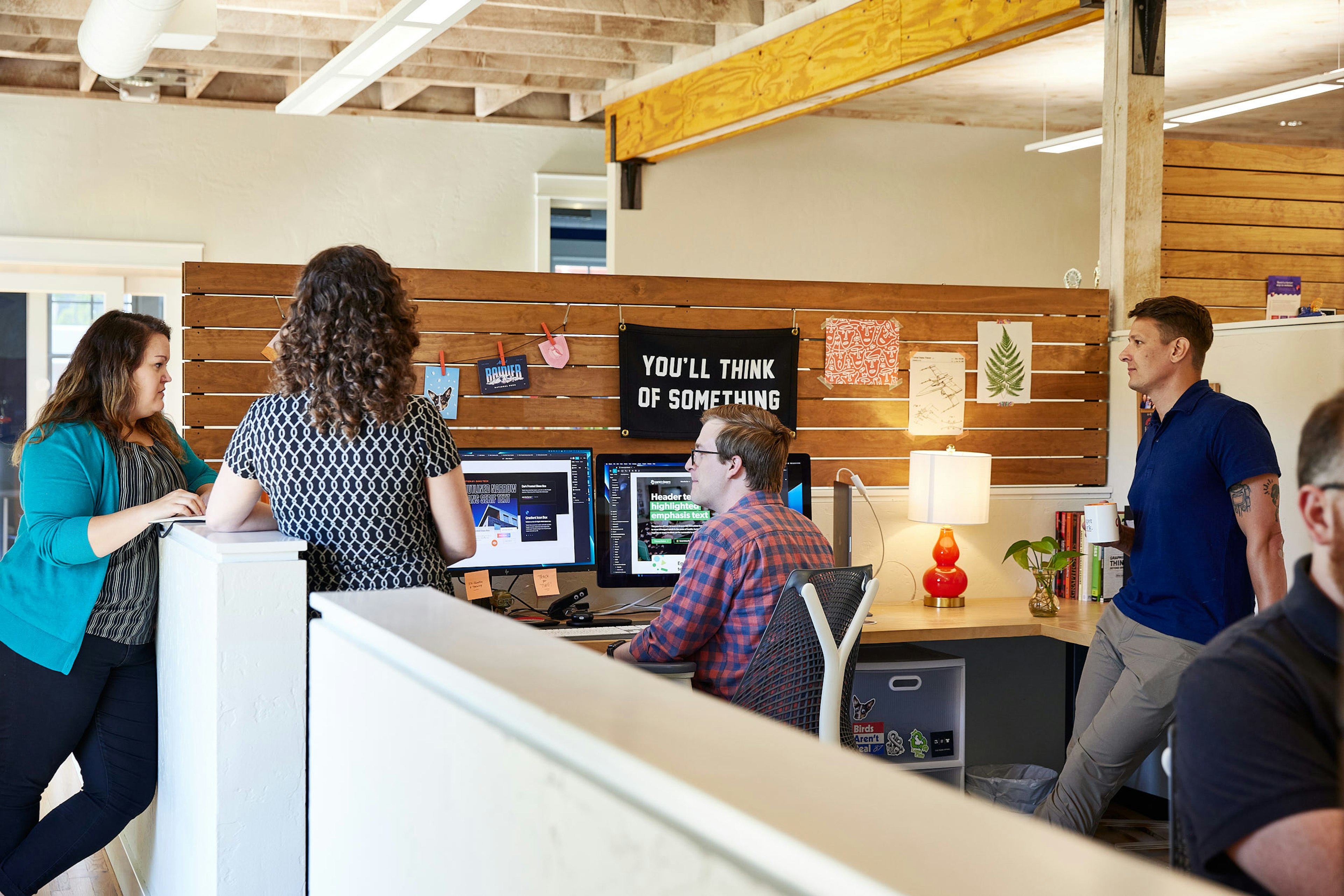 Mostly Serious team members meeting near a desk with dual monitors in an office.