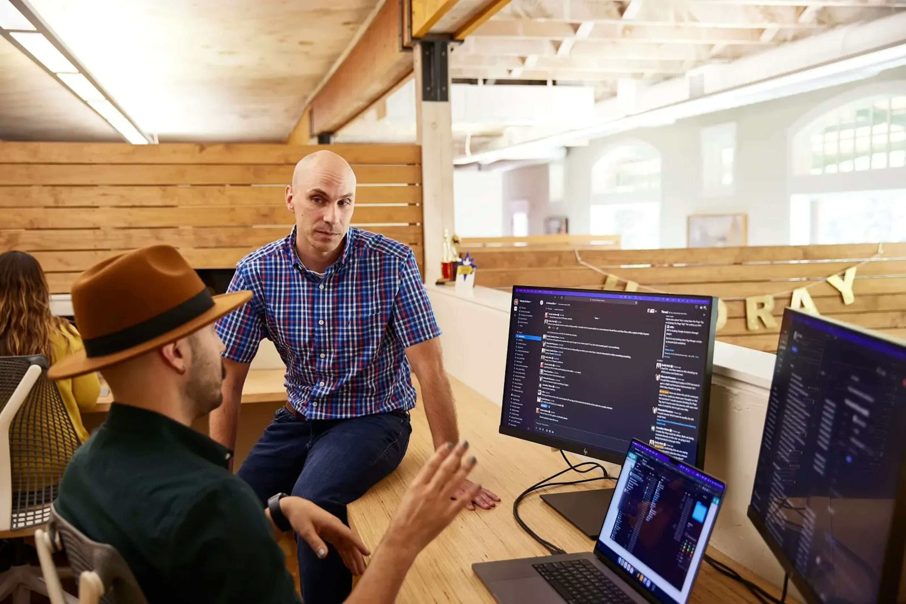 Two Mostly Serious team members collaborating at a shared desk with multiple monitors in a bright, open office.